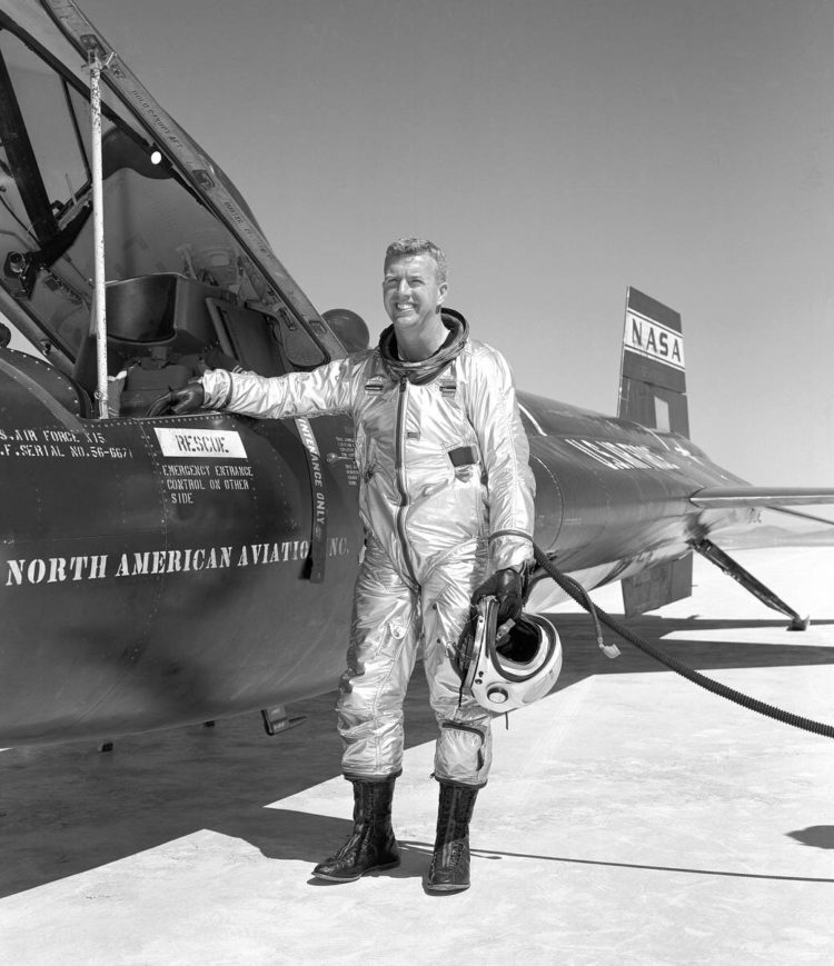 Uśmiechnięty mężczyzna w srebrnym kombinezonie i z hełmem w dłoni stoi obok samolotu rakietowego X-15. 
