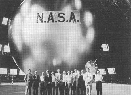 Duży balon o metalicznej powierzchni i napisie N.A.S.A. znajdujący się w hangarze. Przed nim stoi grupa kilkunastu ludzi