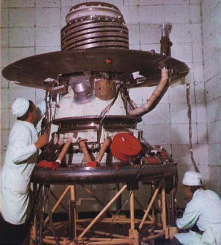 Dwóch naukowców w strojach ochronnych pracujących przy sondzie. Zdjęcie kolorowe.