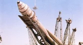 Po środku duża rakieta 3KA, przechylona w kierunku kamery, podnoszona przez metalową konstrukcję od spodu, za nią kilka podobnych metalowych rusztowań, w tle niebo