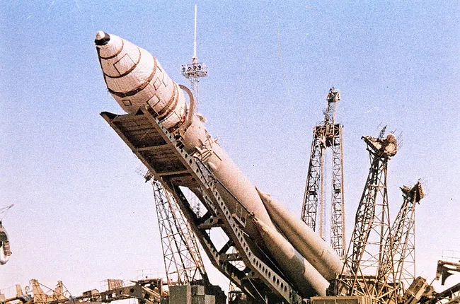 Po środku duża rakieta 3KA, przechylona w kierunku kamery, podnoszona przez metalową konstrukcję od spodu, za nią kilka podobnych metalowych rusztowań, w tle niebo