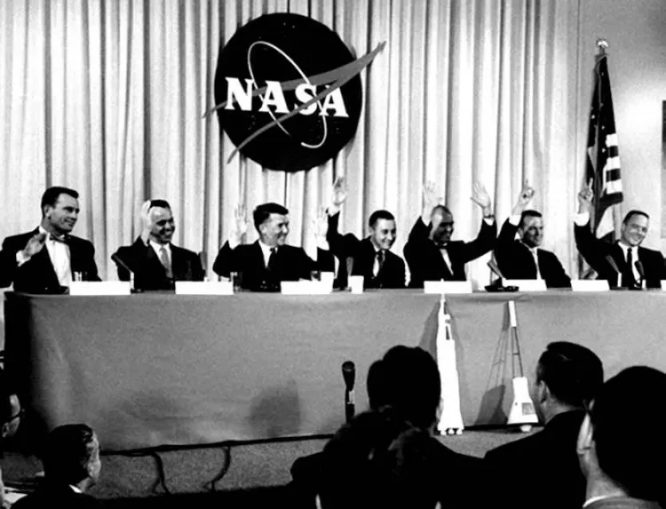 Astronauci programu Mercury (od lewej): Slayton, Shepard, Shirra, Grissom, Glenn, Cooper oraz Carpenter. Po pytaniu, czy czują się pewni, że wrócą na Ziemię po swojej misji, w odpowiedzi wszyscy podnoszą rękę (Glenn podnosi obie). W tle na zasłonie logo NASA.