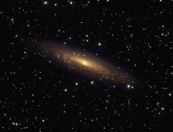 Zdjęcie głębokiego nieba. Przechylona galaktyka w brązowych kolorach ze złotym centrum. W tle liczne gwiazdy.