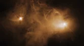 Jasna mgławica emisyjna (obłok gazu i pyłu) rozświetlona na żółto, po lewej i prawej stronie z chmury wyłaniają się dwa jasne punkty, będące gwiazdami