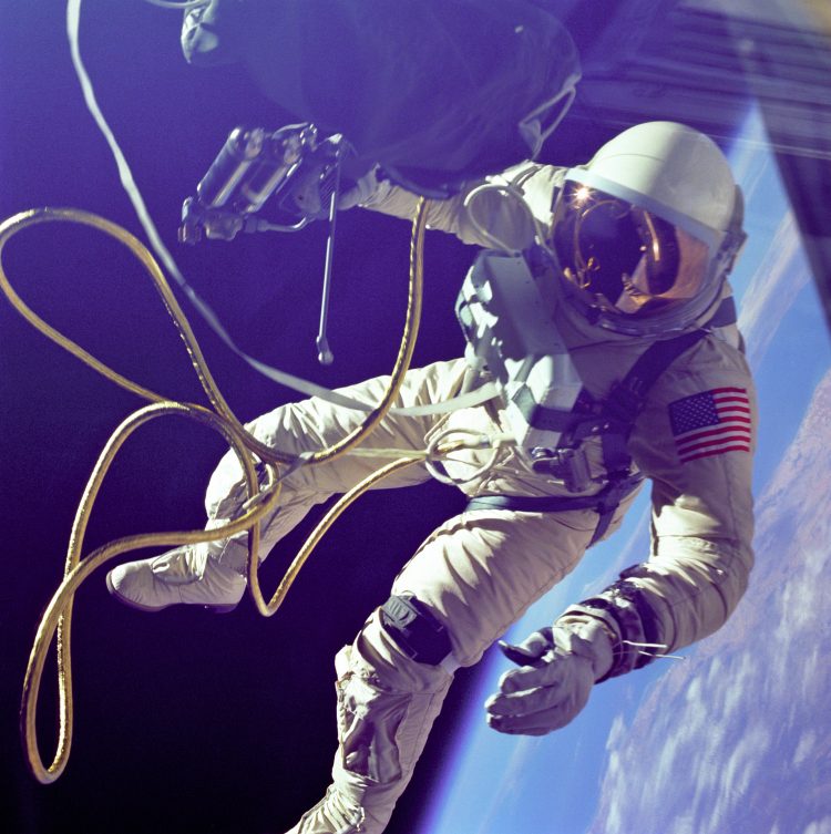 Astronauta w białym skafandrze z naszytą flagą USA na lewym ramieniu. Jest podłączony ze statkiem za pomocą węża, który zwija się przed nim. W tle czarne niebo nad błękitną Ziemią.