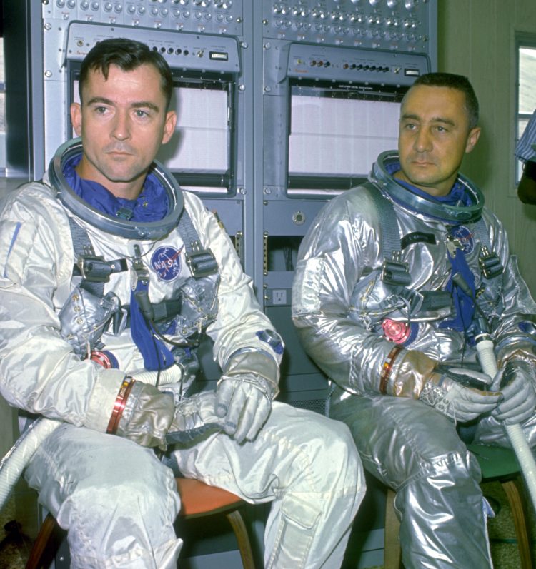 John Young oraz Gus Grissom siedzący w srebrnych skafandrach kosmicznych niezakrywających tylko ich głów. 