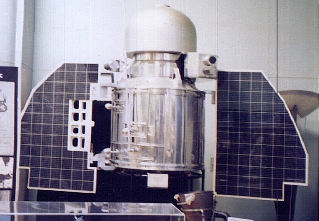 Stojąca w pomieszczeniu sonda złożona ze srebrnego cylindra z białą półkulą na szczycie i dwoma panelami słonecznymi po bokach