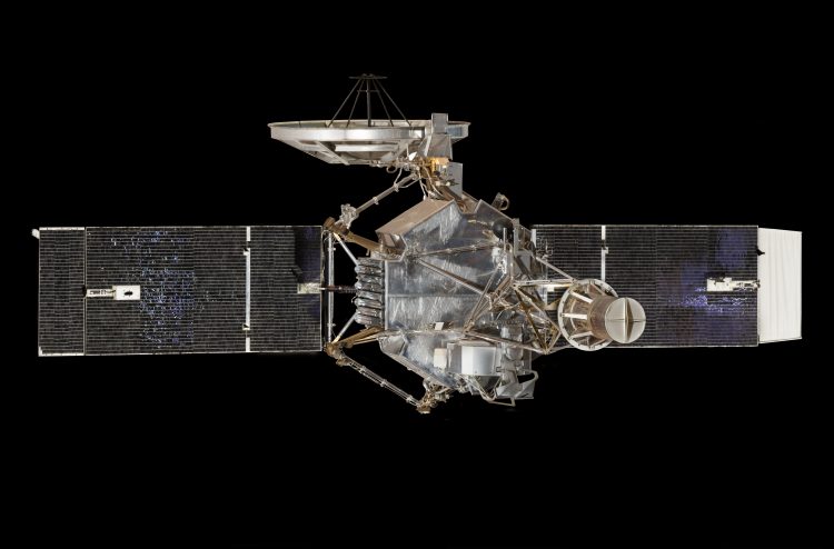Sonda kosmiczna z rozłożonymi dwoma panelami słonecznymi i anteną, o sześciokątnym korpusie i masztem wysuniętym w kierunku aparatu. Tło jest czarne.