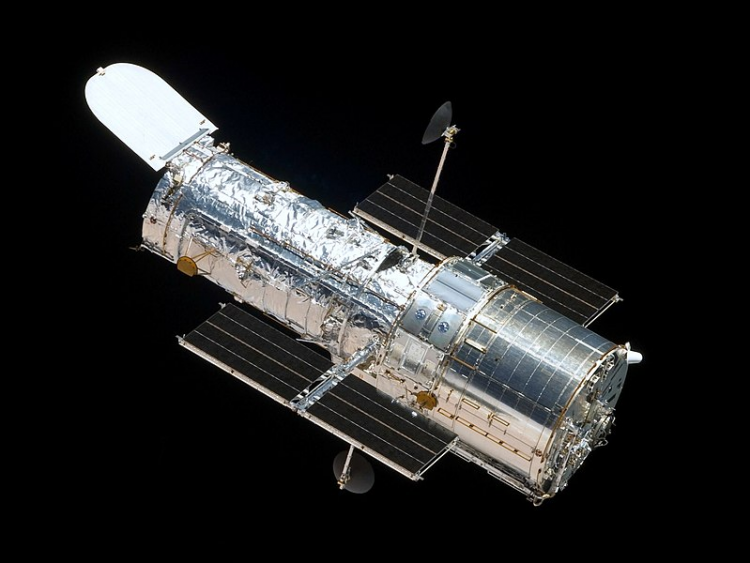 Teleskop Hubble'a, czyli szara metalowa tuba otoczona w górnej części folią izolacyjną, z wystającymi po bokach panelami słonecznymi, na tle czarnego kosmosu.
