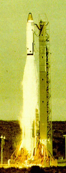 Spiczasta biała rakieta, z czarnym czubkiem, znajduje się kilkadziesiąt metrów nad ziemią, widziana z boku. Z jej spodu wychodzi słup ognia, a obok niej znajduje się metalowa wieża startowa. 