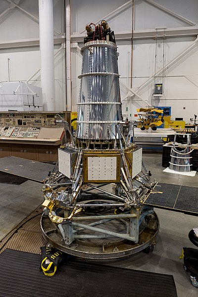 Srebrny satelita w kształcie ściętego stożka stojący w hangarze na platformie na kółkach