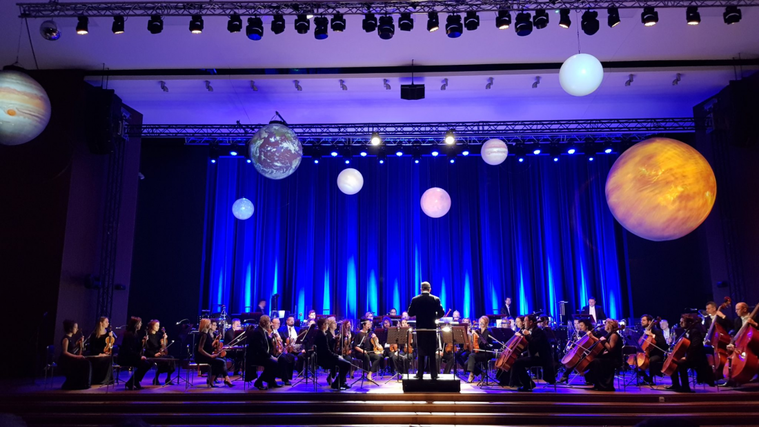 Orkiestra symfoniczna na scenie udekorowanej planetami Układu Słonecznego, które zwisają z sufitu.