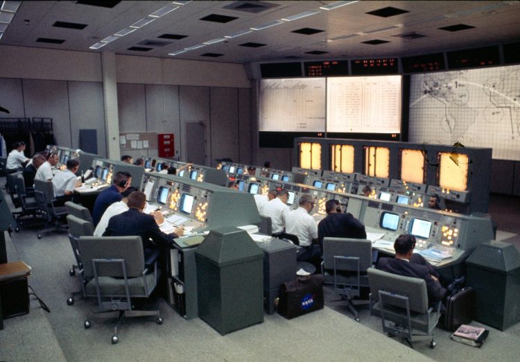 Szare pomieszczenie z dużą ilością komputerów i mężczyznami siedzącymi przy nich; przed nimi na jednej ze ścian jest mapa świata oraz jakieś dane