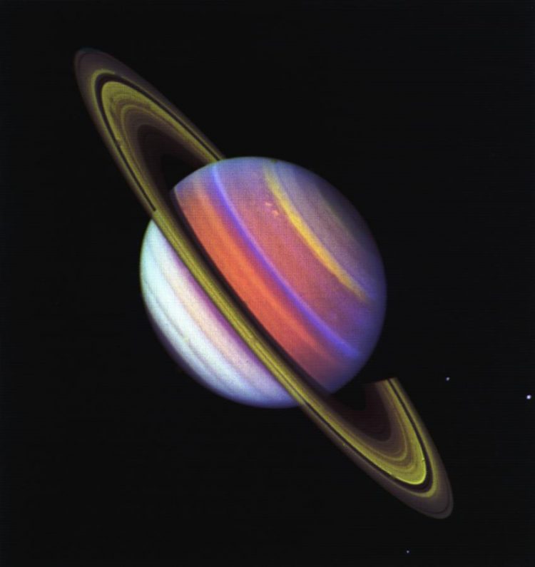 Zdjęcie Saturna ustawionego na ukos. Planeta zamiast beżowego koloru, który widać w świetle widzialnym, jest w fioletowe, żółte, czerwone oraz niebieskie pasy.