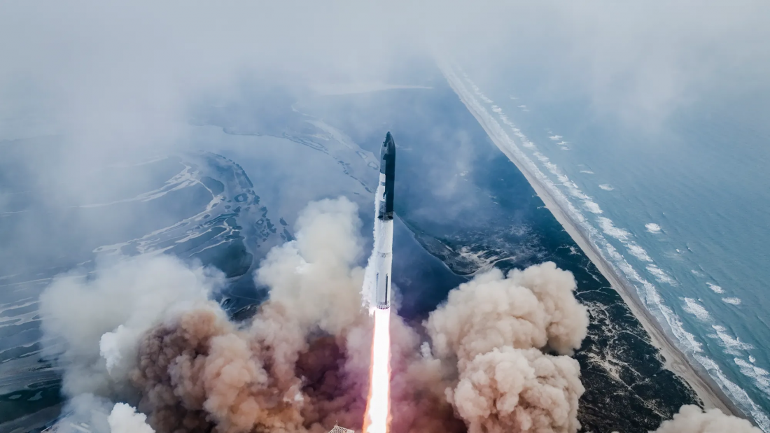 Dwustopniowa rakieta, składająca się z czarnego stopnia Starship i białego stopnia Super Heavy leci w górę. Spod niej wydobywa się potężny słup ognia, który wzbudza ogromne chmury piasku.