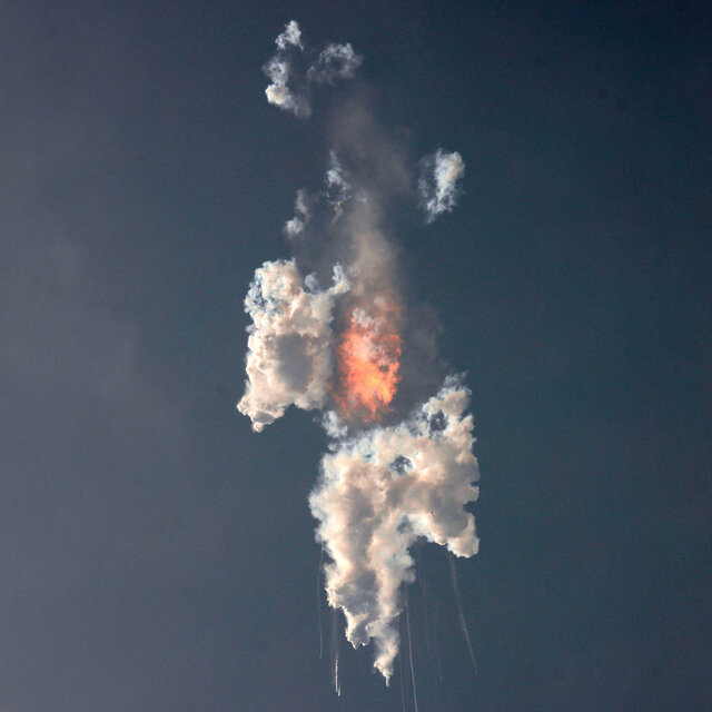 Na niebieskim tle nieba widać ogień, małe, spadające elementy rakiety oraz białe chmury, pozostałe po ciekłym paliwie rakiety 
