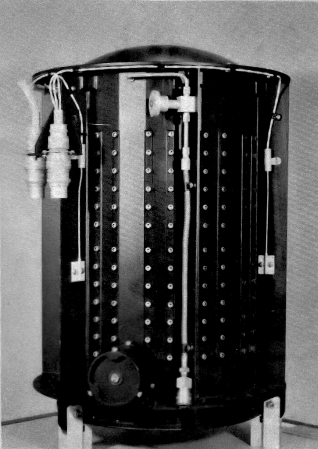Mały cylindryczny metalowy generator z podłączonymi na zewnątrz kablami na tle ściany.