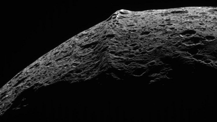 Na pierwszym planie szara Rea z pierścieniami składającymi się z małych szarych kamyków. W tle natomiast niebieska przestrzeń kosmiczna i Saturn z własnymi pierścieniami.