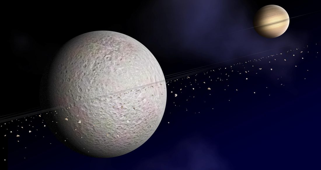 Na pierwszym planie szara Rea z pierścieniami składającymi się z małych szarych kamyków. W tle natomiast niebieska przestrzeń kosmiczna i Saturn z własnymi pierścieniami.
