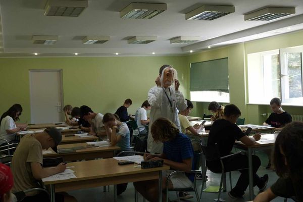 Uczesnicy przy stołach rozwiązują zadania. Pomiędzy nimi stoi kadro w masce Einsteina.