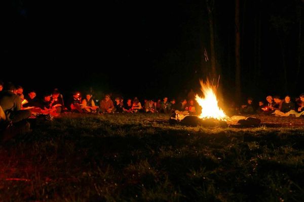 Uczestnicy siedzą na trawie dookoła ogniska, oświetleni pomarańczowym światłem ognia.