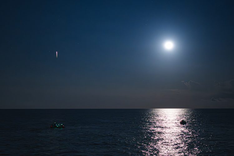 Na tle bezchmurnego nocnego nieba widać jasną smugę, a pod nią oświetlone przez Księżyc w pełni morze i różnokolorowe światła statków odzyskujących.