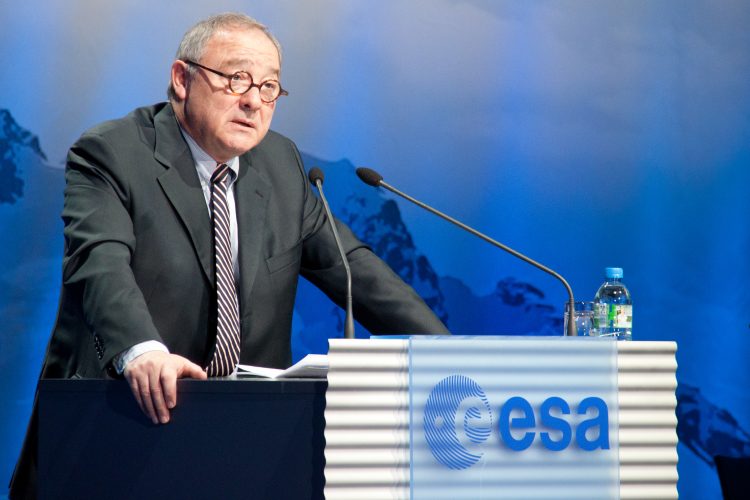 Starszy mężczyzna w okrąglych okularach w garniturze i krawacie stoi za mównicą z dwoma mikrofonami. Mównica jest oznaczona logo ESA, w tle znajduje się zdjęcie gór.