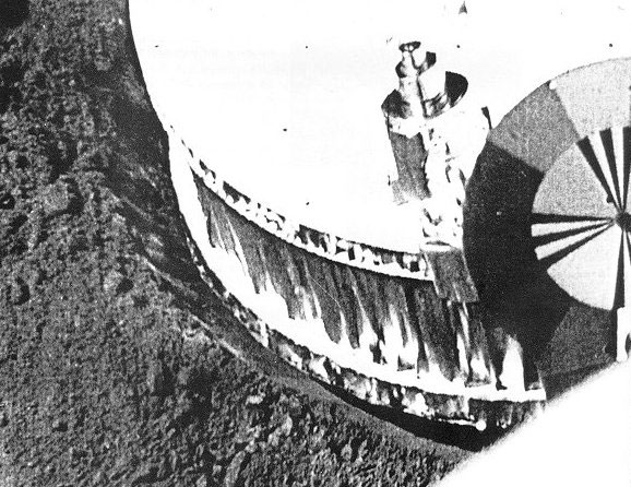 A black and white picture of a cylindrical white object, which happens to be the footpad of the probe. It created a small creater in a sand like material, being the Moon regolith.