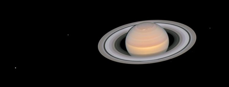 Na czarnym tle po prawej widnieje pomarańczowo kremowa kula, na której widać pasy. Jest ona okolona pierścieniami. Ta kula to Saturn. W te widać nieliczne białe kropki - gwiazdy.