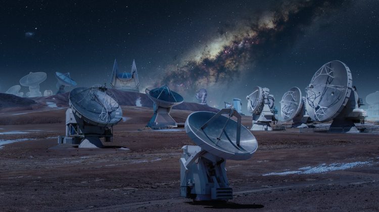 Na brązowym wzgórzu liczne szare teleskopy radiowe, skierowane w różne strony. W tle widoczna Droga Mleczna na nocnym niebie.