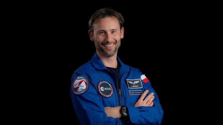 Zdjęcie przedstawia polskiego astronautę Sławosza Uznańskiego-Wiśniewskiego, stojącego na czarnym tle w niebieskim uniformie z naszywkami ESA, flagą Polski oraz swoim nazwiskiem.