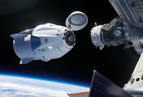 Na ilustracji przedstawiono kapsułę kosmiczną Crew Dragon firmy SpaceX w trakcie zbliżania się do portu dokującego Międzynarodowej Stacji Kosmicznej. Pokrywa portu dokowania kapsuły jest otwarta. Widoczna jest Ziemia w tle oraz fragment paneli słonecznych stacji. Na kapsule znajdują się oznaczenia NASA, SpaceX oraz flaga USA.