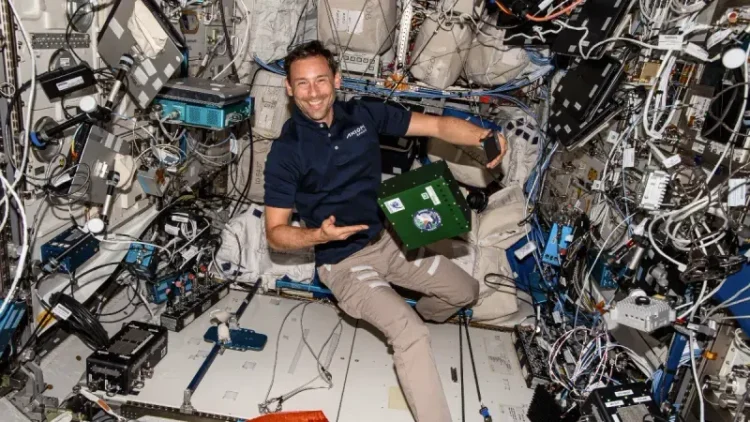 Astronauta w granatowej koszulce z kołnierzykiem i beżowych spodniach, o ciemnych krótkich włosach i lekkim zaroście wskazuje na lewitującą, zieloną skrzynkę. Cała scena dzieje się w otoczeniu kabli, pudełek i monitorów Międzynarodowej Stacji Kosmicznej. 