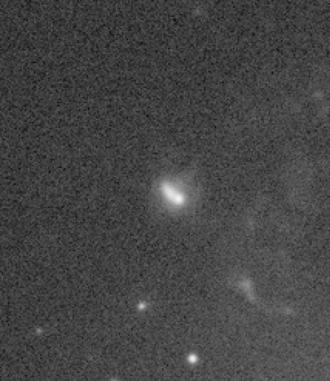 Powiększony wycinek surowego obrazu astronomicznego z wyraźnym, drobnoziarnistym szumem luminancji; pośrodku widać niewielką, lekko pochyloną galaktykę, a w tle liczne jasne i ciemne piksele tworzą gęstą ziarninę.