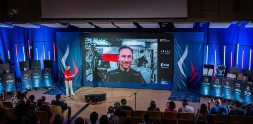 W tle widać duży baner na którym wyświetla się postać mężczyzny o krótkich ciemnych włosach i lekkim zaroście. Po lewej stronie na scenie stoi kobieta w czerwonej koszulce oraz białych spodniach. Przy dolnej krawędzi zdjęcia widać ludzi siedzących przed sceną.