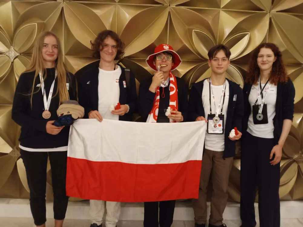 Pięć osób stojących na tle złotej ściany pokrytej wypukłym wzorem w kształcie kwiatów. Trzymają rozłożoną flagę Polski