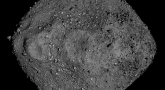 Obraz przedstawia asteroidę Bennu, sfotografowaną z bliska. Ma ona nieregularny, niemal diamentowy kształt i pokryta jest luźnymi skałami oraz pyłem. Powierzchnia wygląda na chropowatą i porowatą, a tło jest całkowicie czarne.