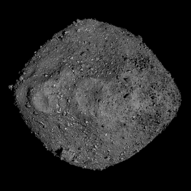 Obraz przedstawia asteroidę Bennu, sfotografowaną z bliska. Ma ona nieregularny, niemal diamentowy kształt i pokryta jest luźnymi skałami oraz pyłem. Powierzchnia wygląda na chropowatą i porowatą, a tło jest całkowicie czarne.