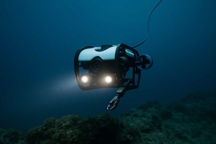 Robot, wyglądający trochę jak nieobudowany samochód ze ramieniem do pobierania próbek, płynie pod wodą nad ciemnozielonymi skałami. Woda jest ciemnogranatowa, robot świeci dwoma światłami, nad którymi znajduje się kamera. 