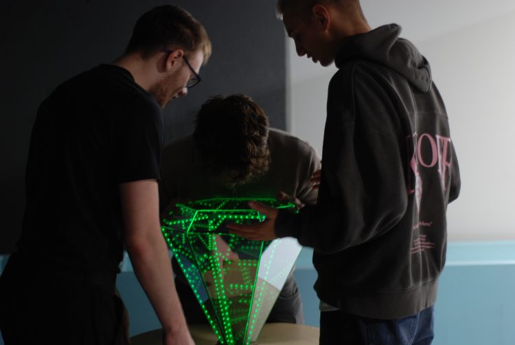 Zdjęcie przedstawia trzech uczestników wydarzenia wokół eksponatu z wystawy w Epi-Centrum Nauki; świecącym na zielono kryształem.