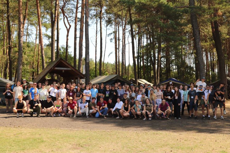 Uczestnicy obozu oraz ich opiekunowie pośród natury i namiotów podczas słonecznego dnia.