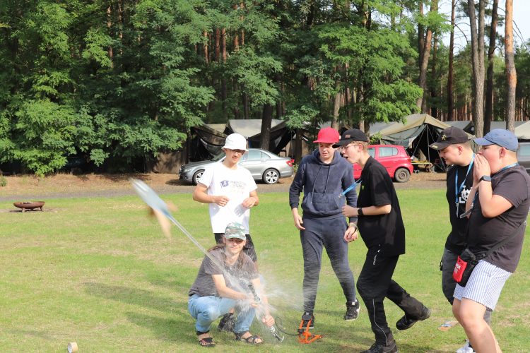 Drużyna młodzieży ekscytująca się wystrzeliwaniem rakiety zbudowanej z plastiku i papieru, w tle znajduje się natura oraz namioty.
