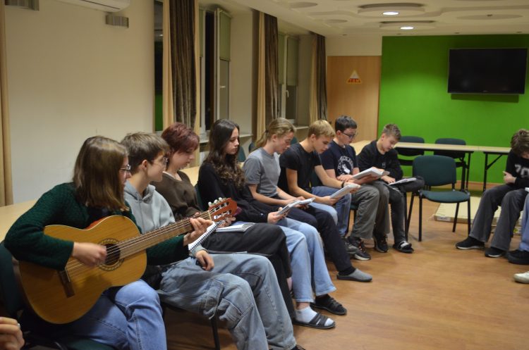 Kilka osób siedzących w rzędzie na krzesłach przy ścianie. Trzymają gitary i śpiewniki.