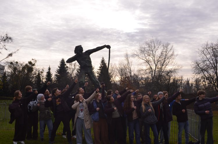 Grupa osób stojących pod pomnikiem napinającego łuk mężczyzny i naśladujących jego pozycję. W tle park.