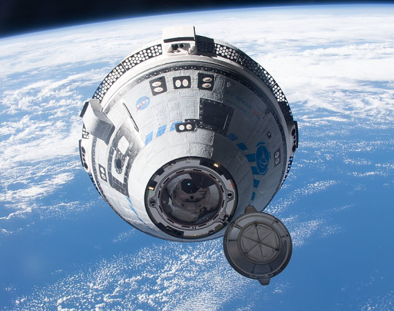 Kapsuła kosmiczna widziana z bliska na orbicie okołoziemskiej, z otwartym włazem dokującym na pierwszym planie; w tle zakrzywiony horyzont Ziemi, błękitny ocean i chmury.