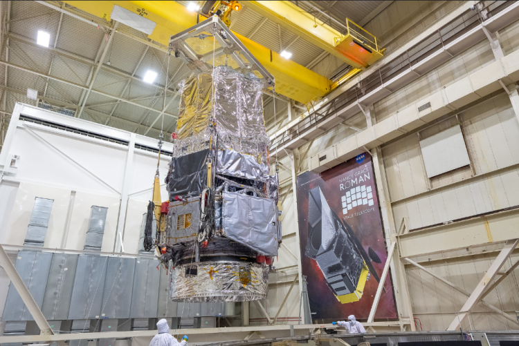 Teleskop Nancy Grace Roman w hali montażowej podczas testów termicznych. Na drugim planie plakat z wizualizacją końcowego efektu.