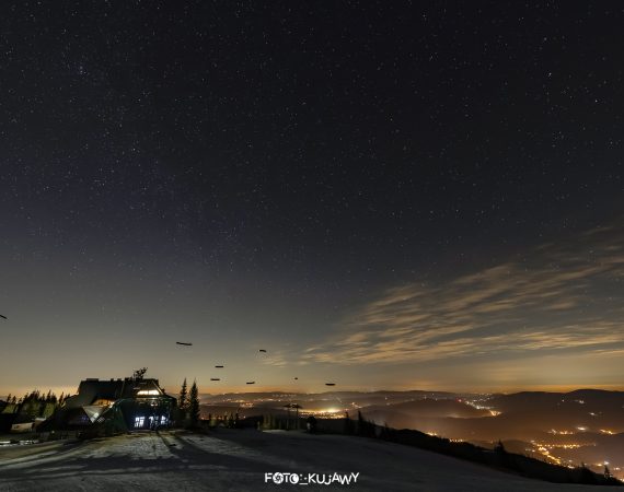 W dolnej części zdjęcia niewysokie góry zimą, w górnej nocne niebo.