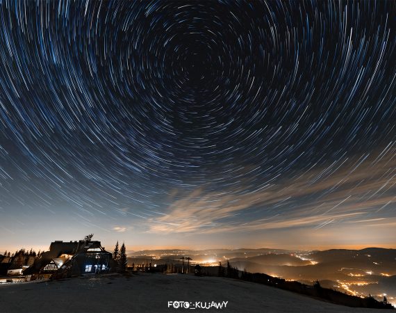Niewysokie góry pokryte śniegiem w dolnej części zdjęcia, na górze gwiazdy ciągnące za sobą smugi i układające się w wir, którego centralny punkt znajduje się w środkowo-górnej części nieba.