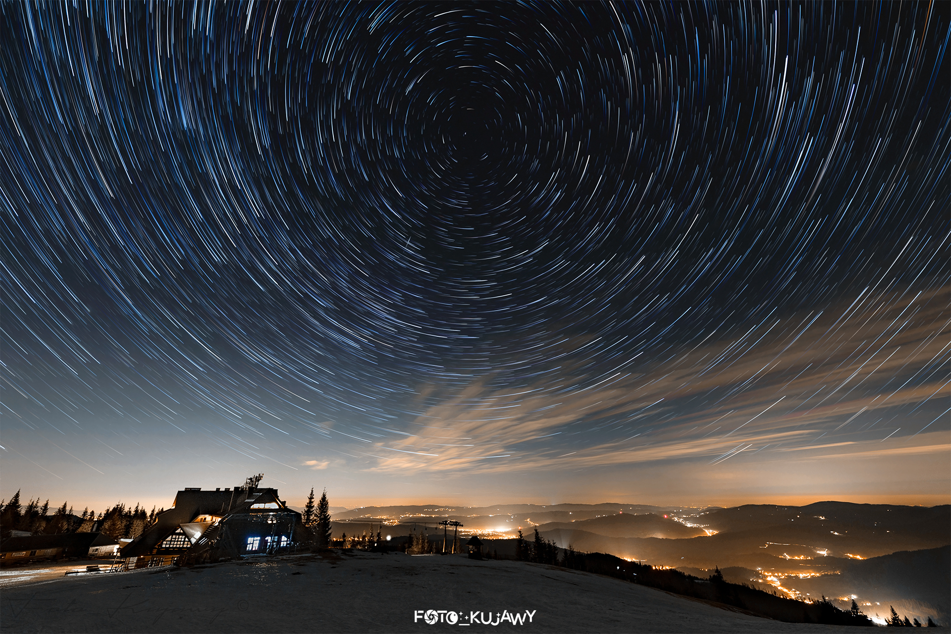 Niewysokie góry pokryte śniegiem w dolnej części zdjęcia, na górze gwiazdy ciągnące za sobą smugi i układające się w wir, którego centralny punkt znajduje się w środkowo-górnej części nieba.