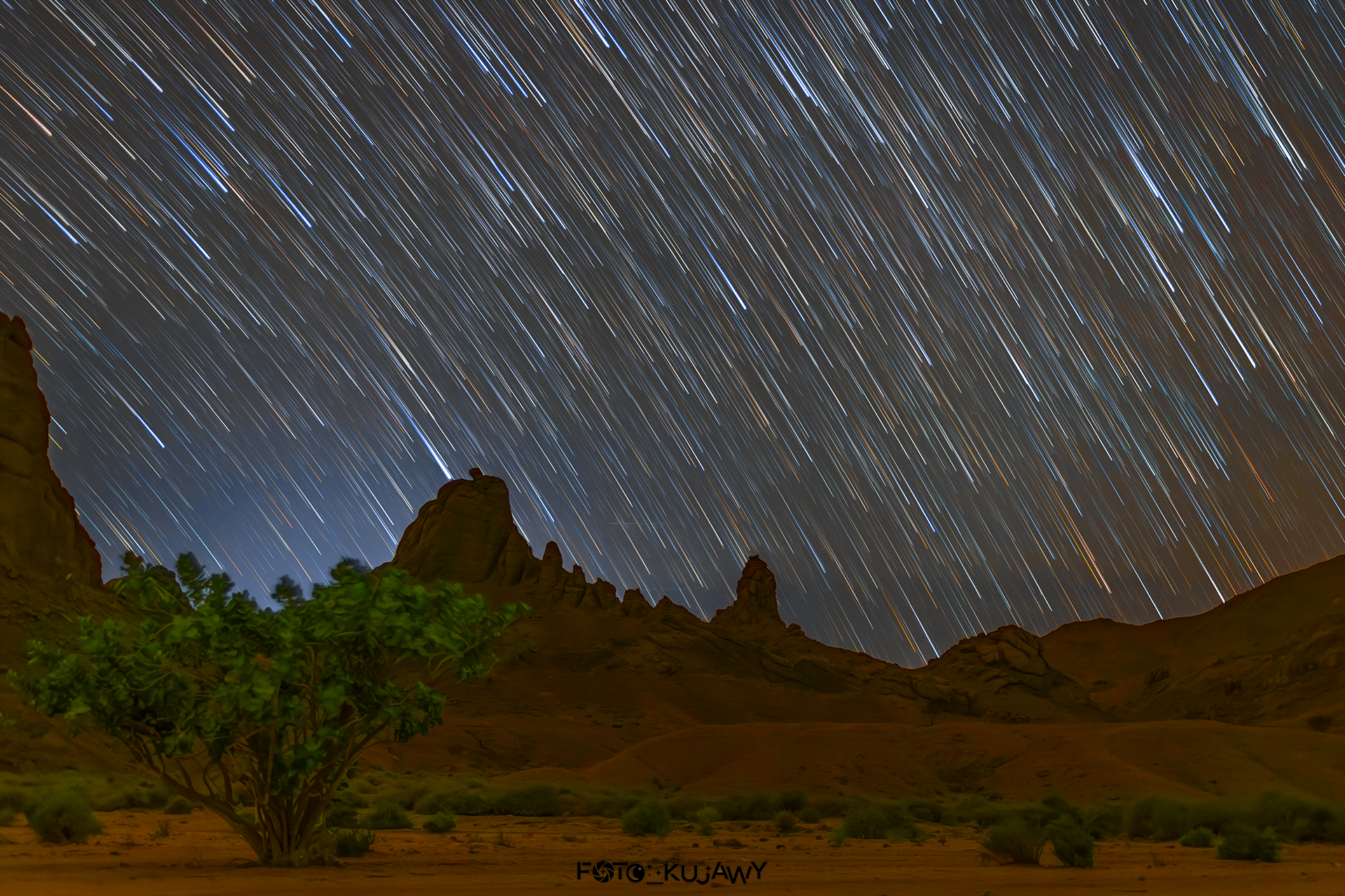 Pustynny pomarańczowy skalny krajobraz w dolnej części zdjęcia, na górze gwiazdy ze smugami za nimi.
